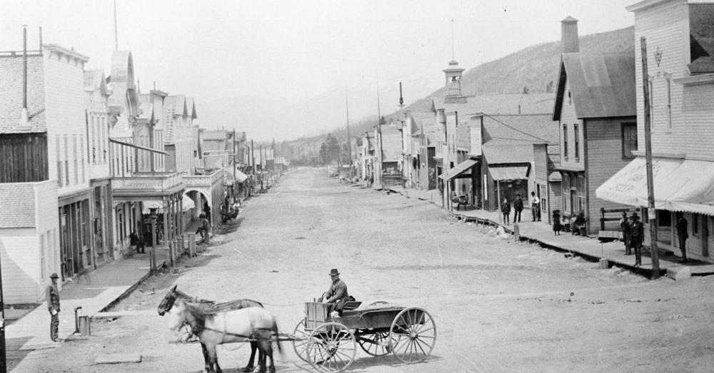 Historic Main Street in Breckenridge, Colorado, circa 1880s -- a horse-drawn wagon on a dirt road lined with wooden storefronts and mountains in the background